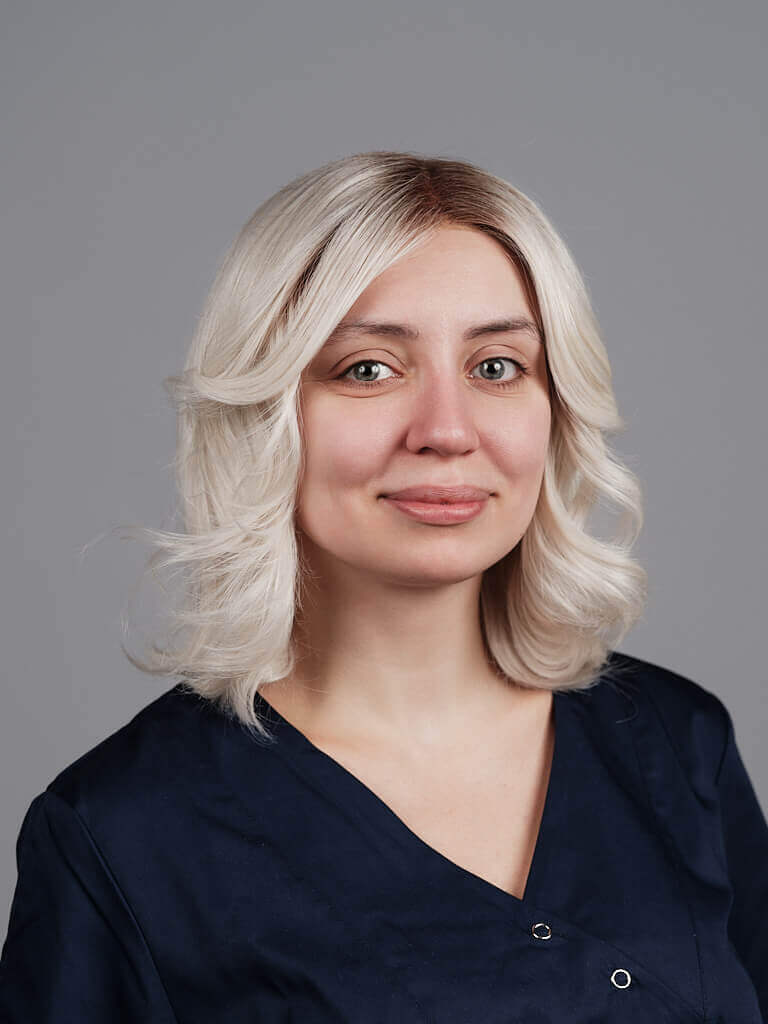 Porträt einer lächelnden blonden Frau mit gewelltem Haar und dunklem Ansatz in einem dunkelblauen Kasack vor grauem Hintergrund.
